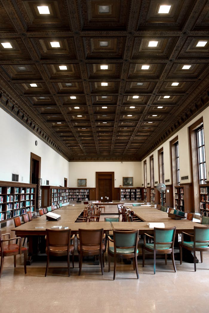 Spacious library with wooden tables and chairs, surrounded by bookcases, illuminated by ceiling lights.
